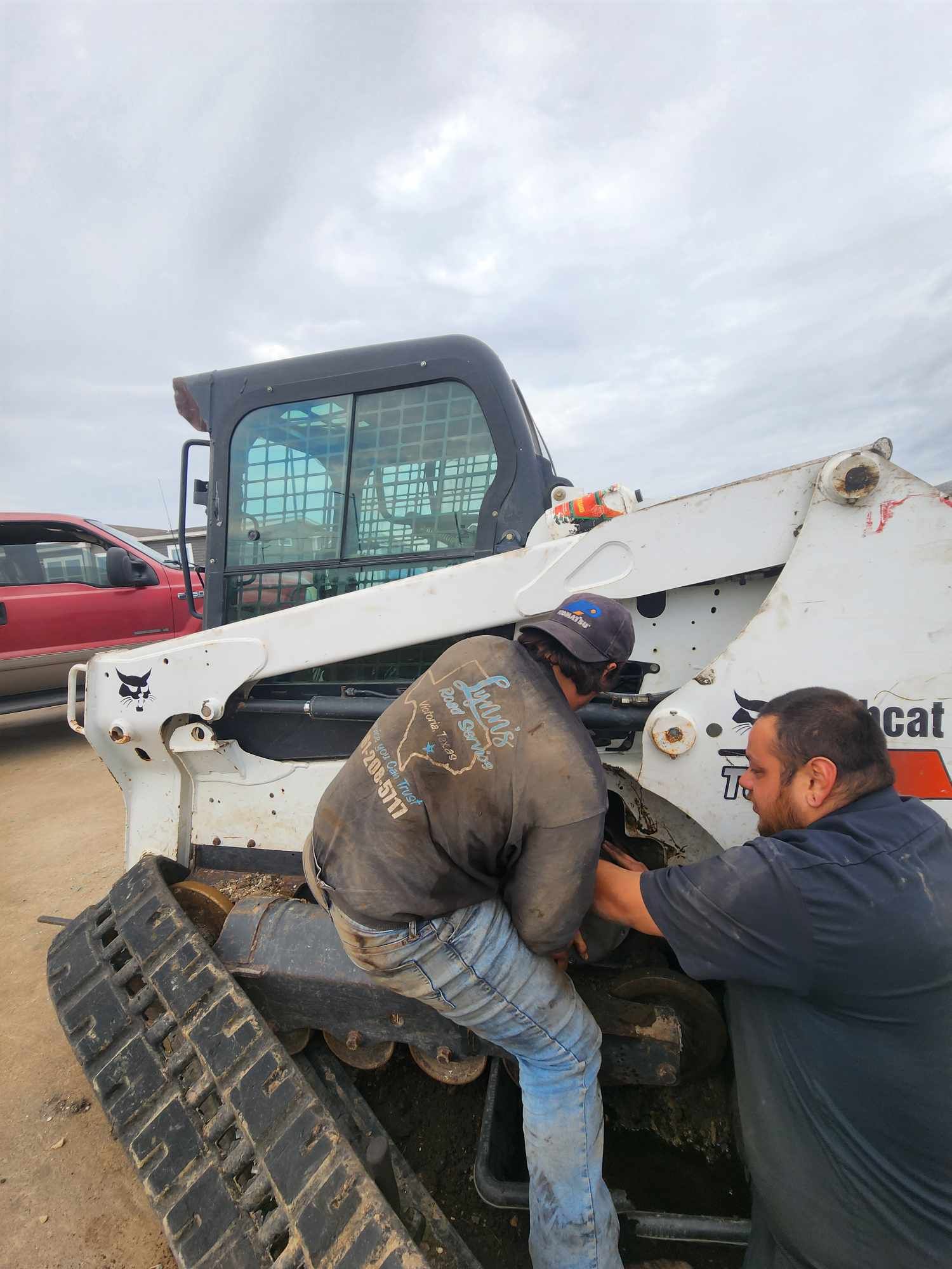 Two men working on a white Bobcat track loader outdoors; cloudy sky.