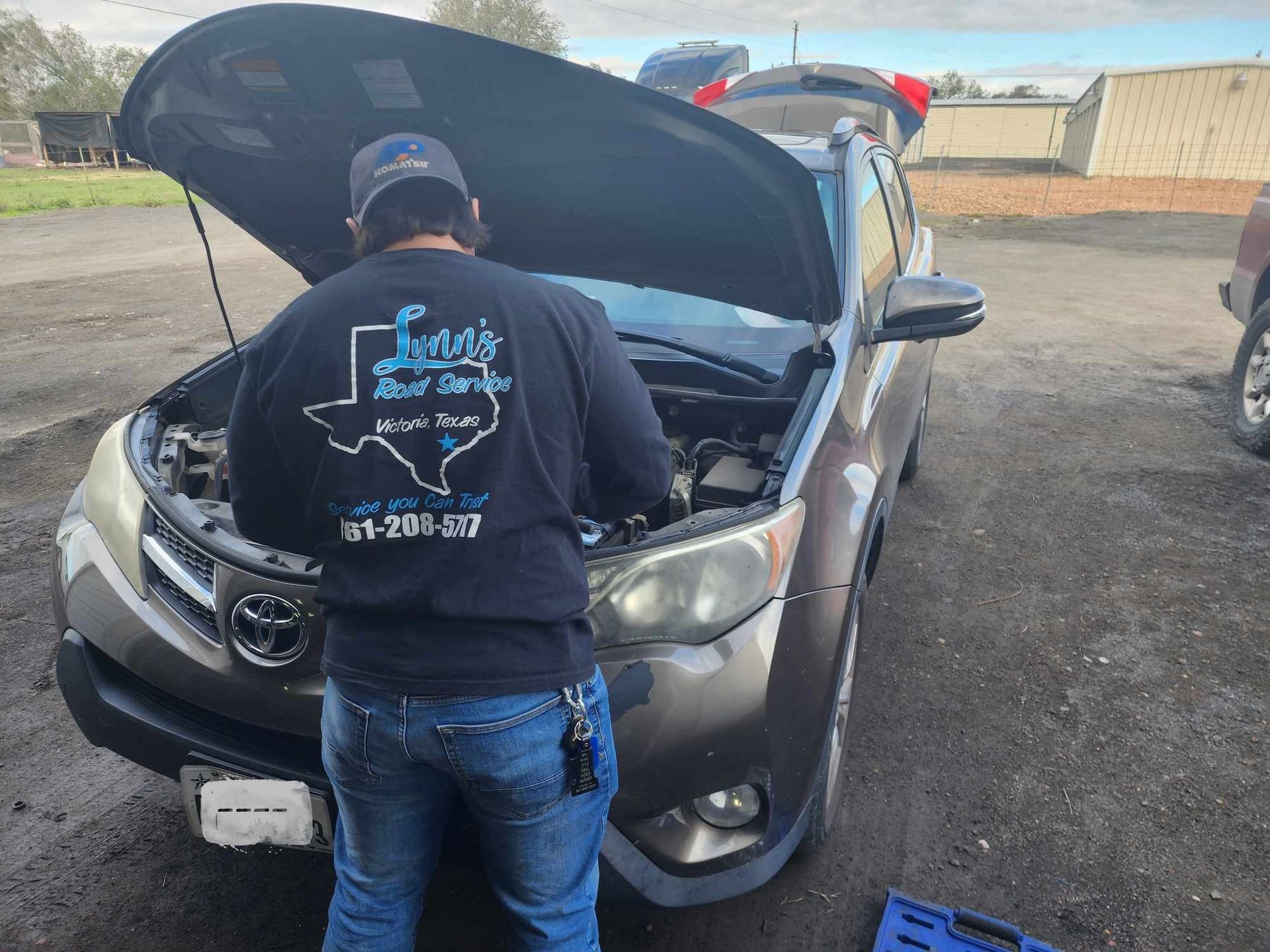 Mechanic working on a brown Toyota SUV with the hood open in an outdoor setting.