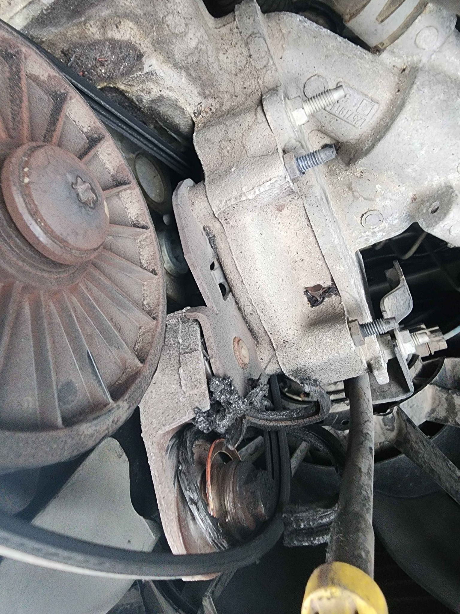 Damaged car alternator with melted components, beside a fan and belt, in an engine compartment.