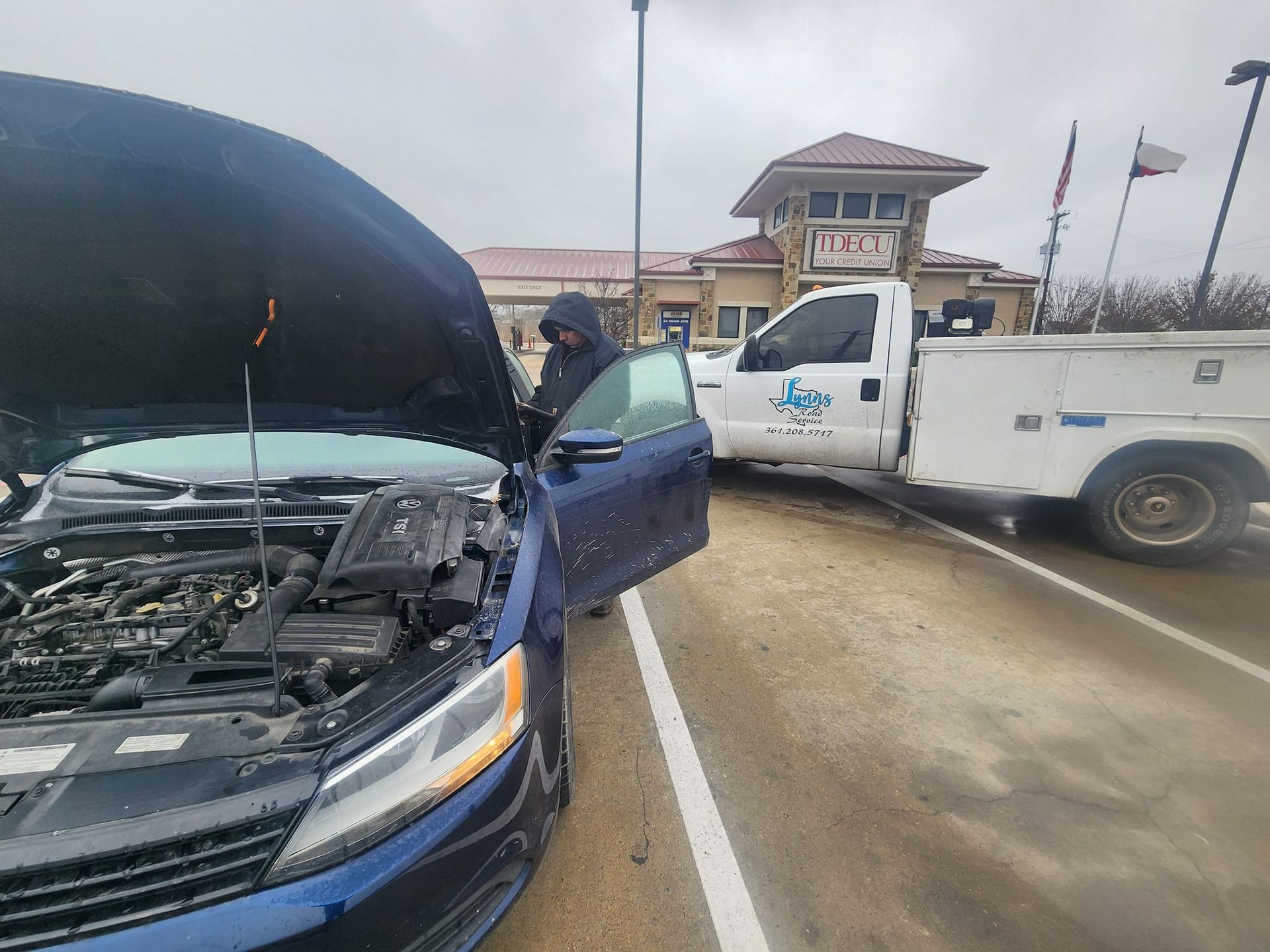 Person by blue car with open hood, tow truck behind in parking lot, overcast day.