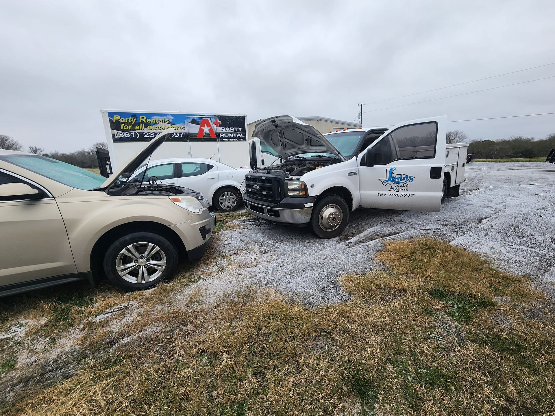 Cars with open hoods, roadside, cloudy day. One has a service truck with logo.