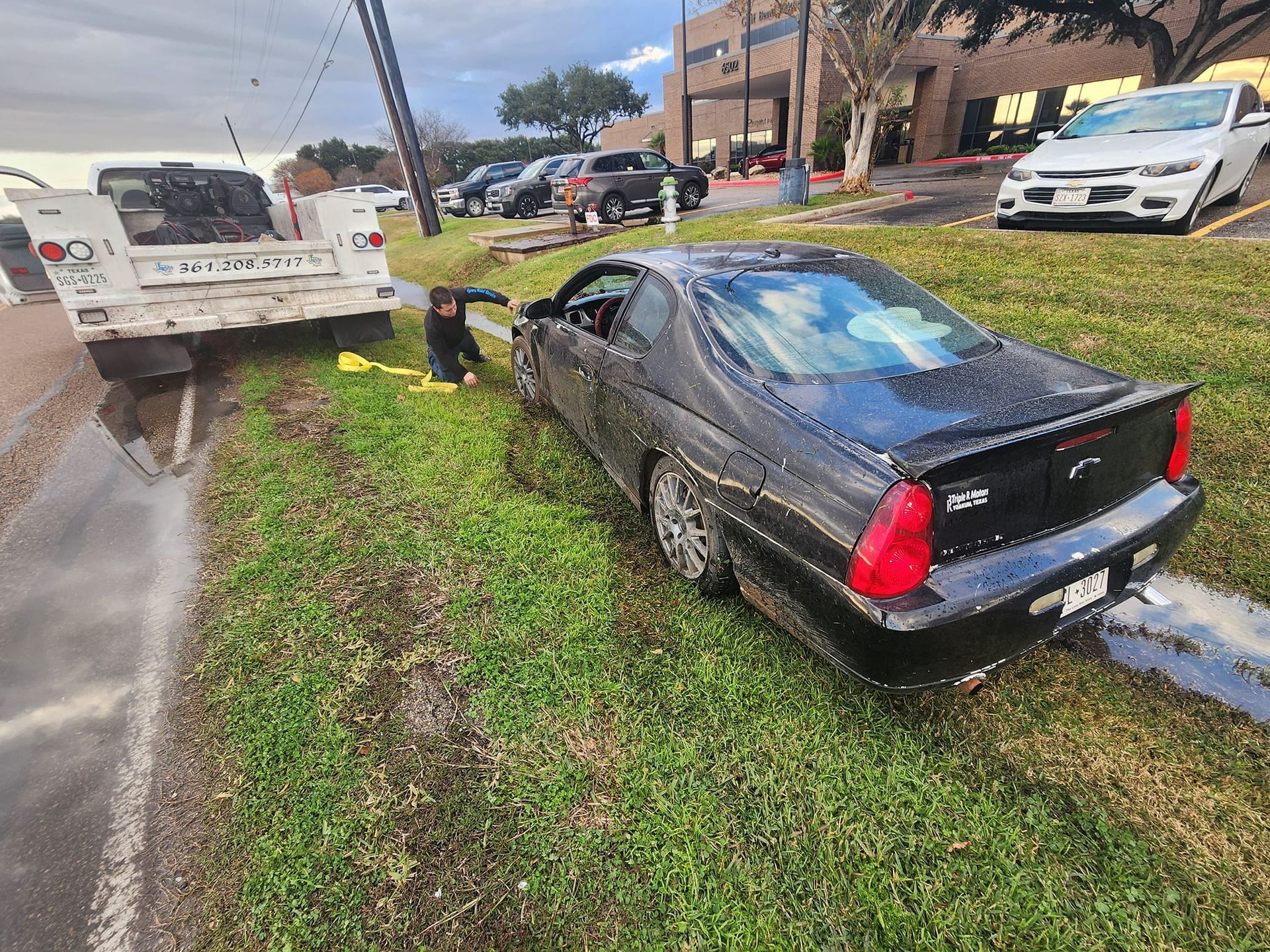Black car stuck in ditch, man assisting. Tow truck nearby, setting is outdoors, daytime.