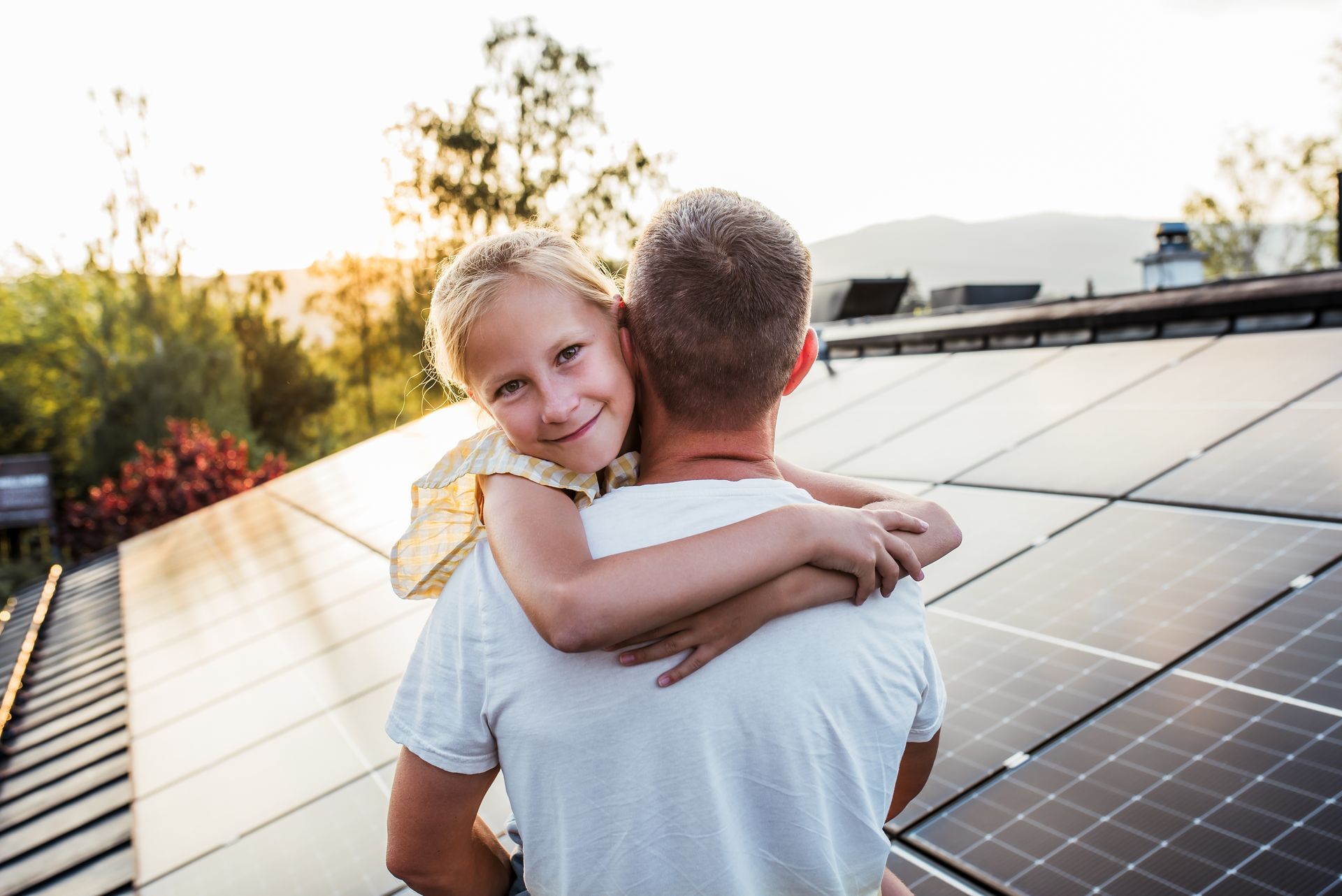 A man is holding a little girl on his shoulders in front of solar panels.