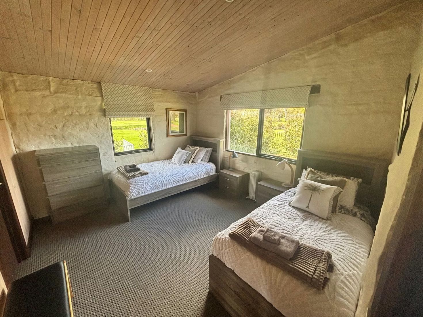 A bedroom with stone walls, two twin beds, patterned carpet, and wooden ceiling, brightened by two windows.