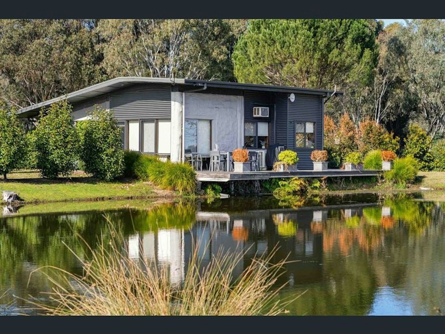 A modern cabin with dark wood siding and a light stone facade sits on the edge of a calm pond reflecting the trees.