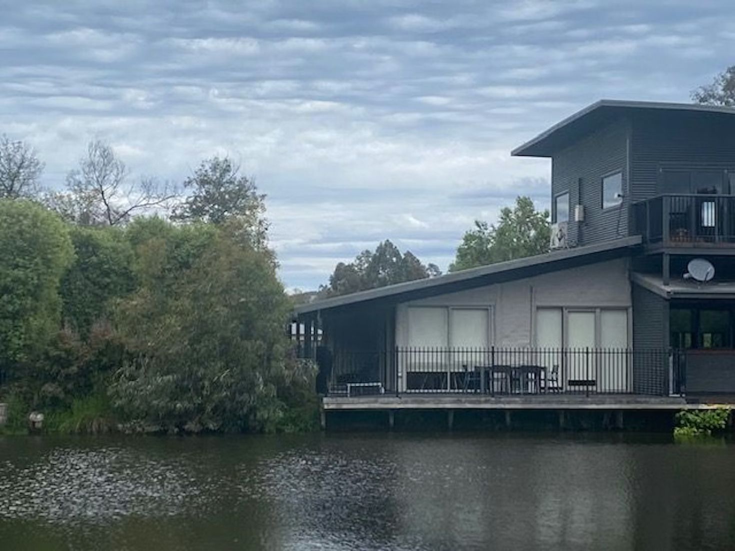 A modern, grey two-story house with a wooden deck overlooking a calm lake surrounded by lush green trees.