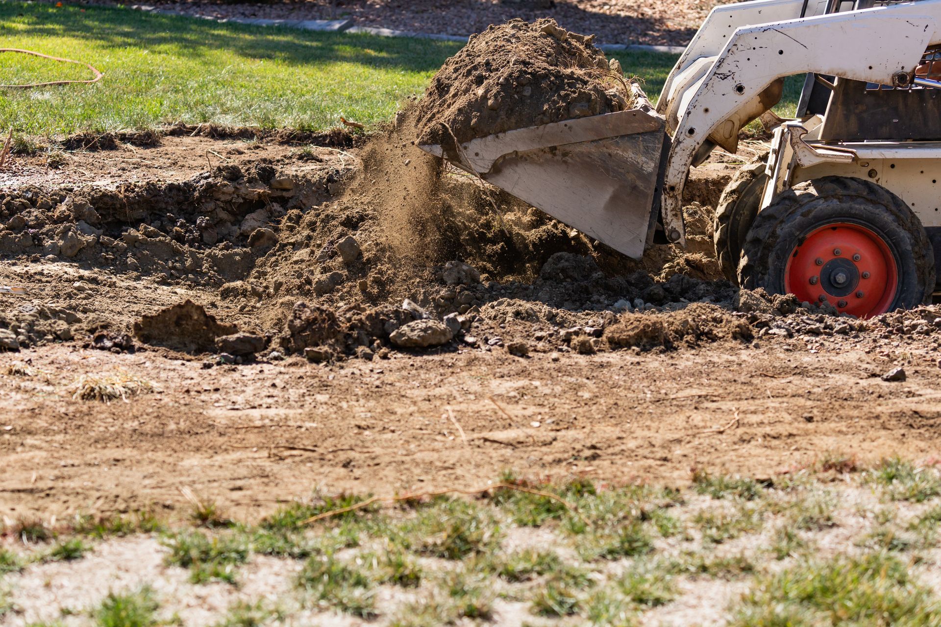 Small skid steer digging and dumping dirt on a grassy, muddy site