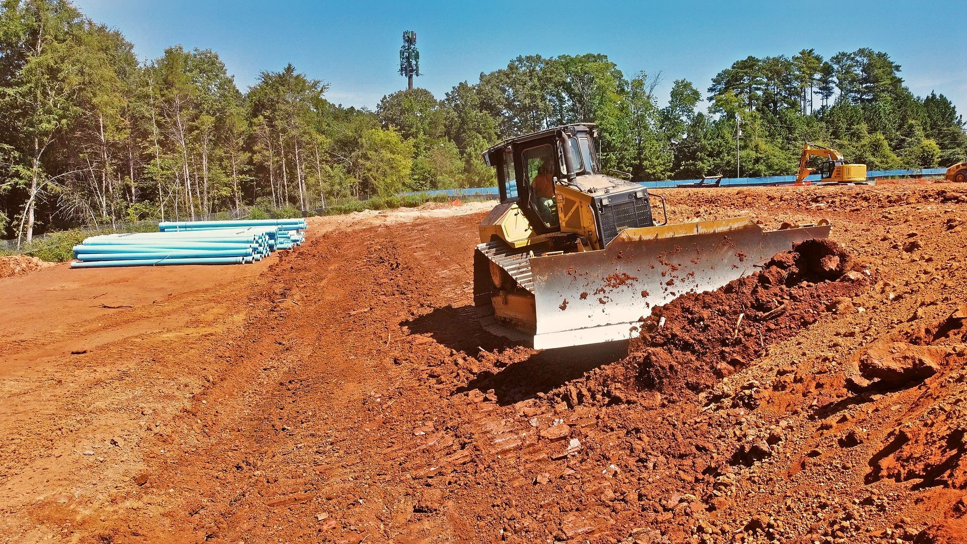 Bulldozer grading a red dirt construction site with stacked blue pipes and trees in the background