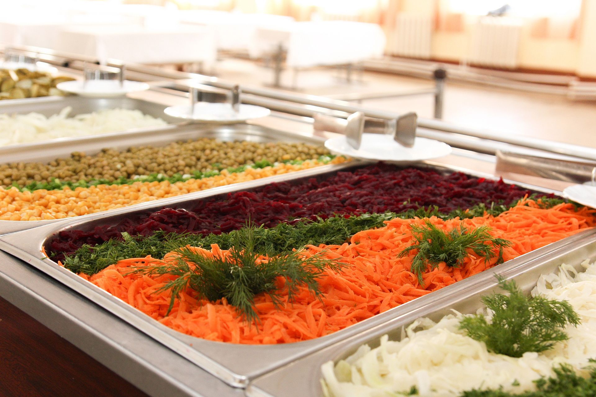 Un tavolo da buffet con vassoi di metallo pieni di file di carote grattugiate, barbabietole, verdure a foglia verde e cipolle bianche.