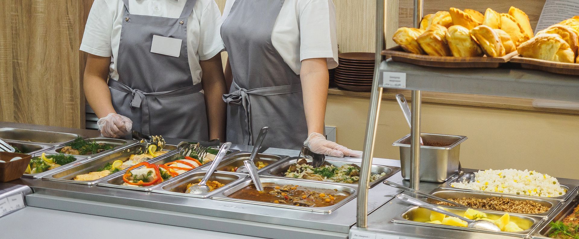 Due persone con grembiuli grigi servono vari piatti caldi da un buffet al banco della ristorazione.