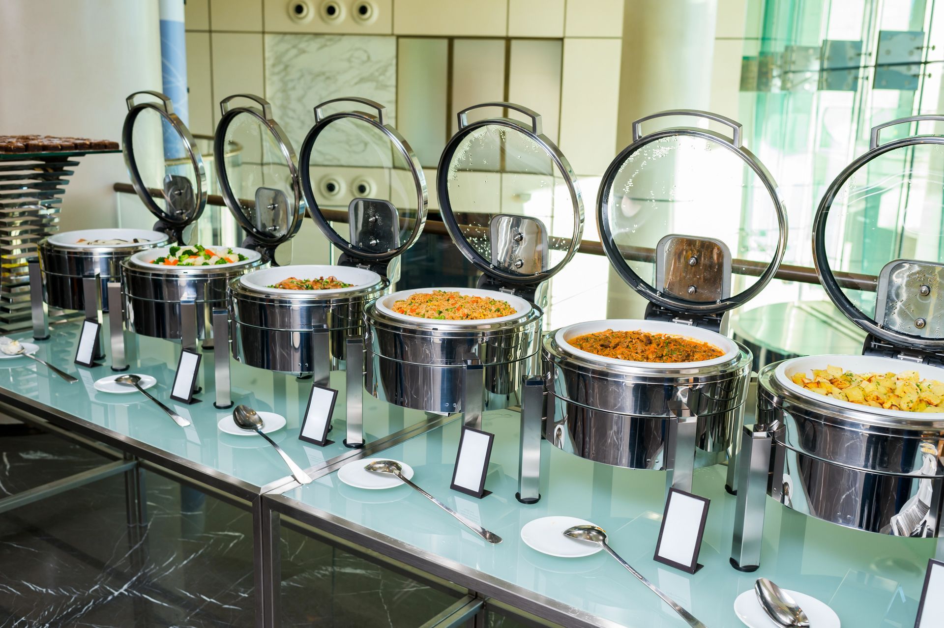 In una sala da pranzo, su un tavolo di vetro, si trova una linea di buffet con diversi scaldavivande in argento contenenti varie pietanze calde.