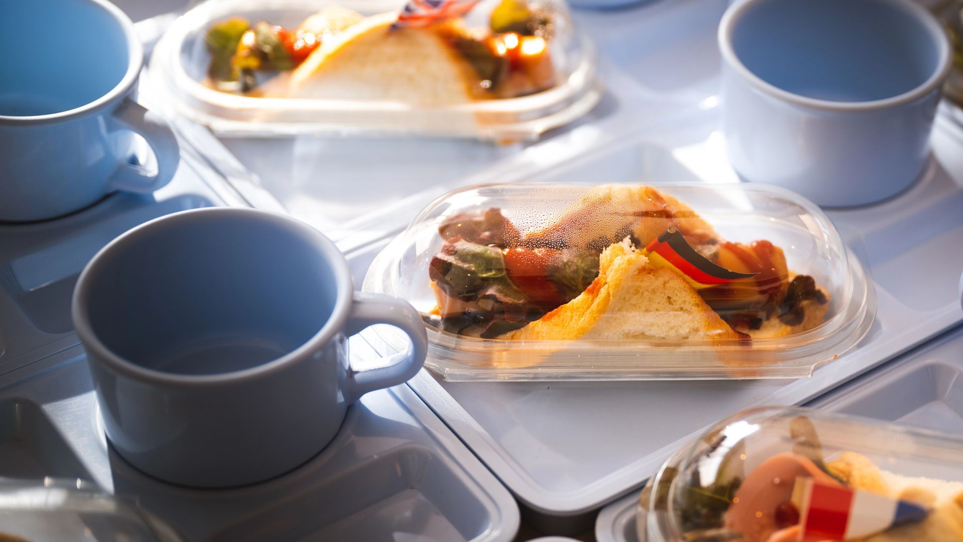 Su un tavolo di colore chiaro sono appoggiati bicchieri di plastica blu e vassoi per alimenti ricoperti di plastica contenenti panini.