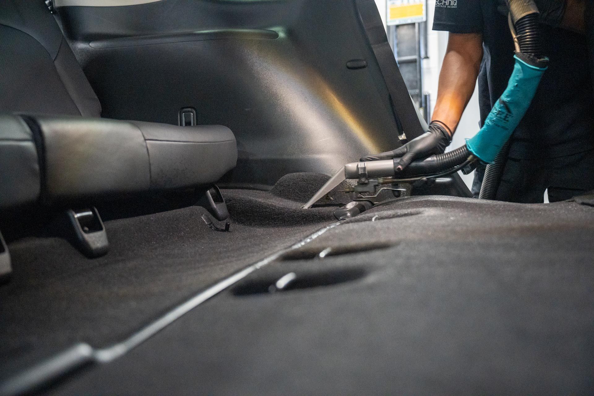 Person vacuuming car interior; black gloves, teal sleeve, dark gray carpet.