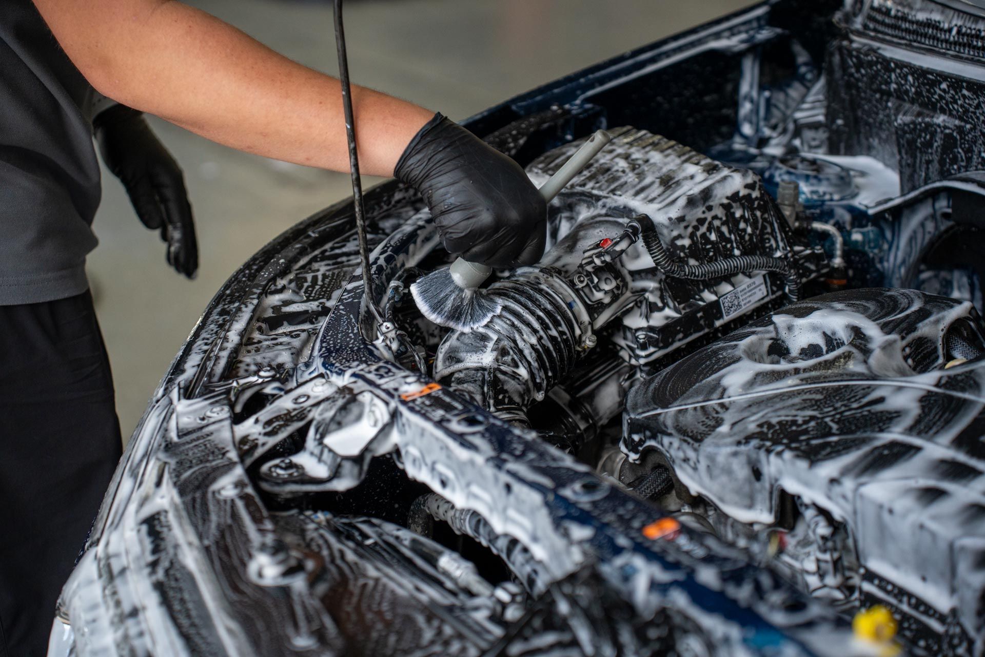 Person in black gloves washing a car engine with foamy cleaner.