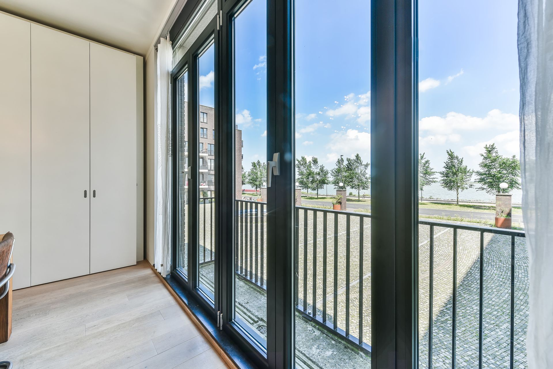 Interior view with large black-framed windows open to a balcony overlooking a brick patio and trees against a blue sky.