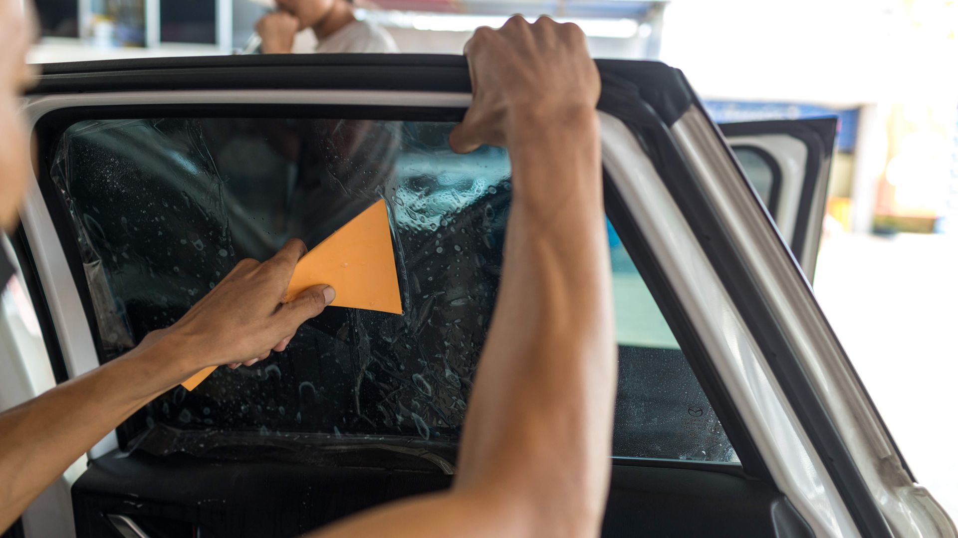 Person applying tint to a car window with a tool, outdoors.