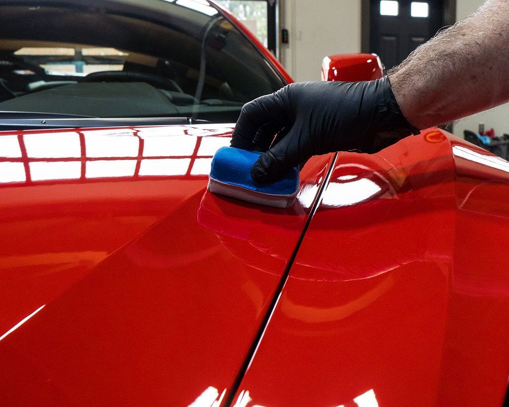 Gloved hand applying a blue sealant block to a shiny red car's body panel. Orange cloth nearby.