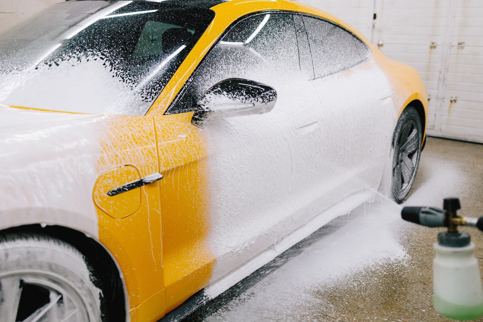 Yellow sports car covered in white foam, being washed in a garage setting.