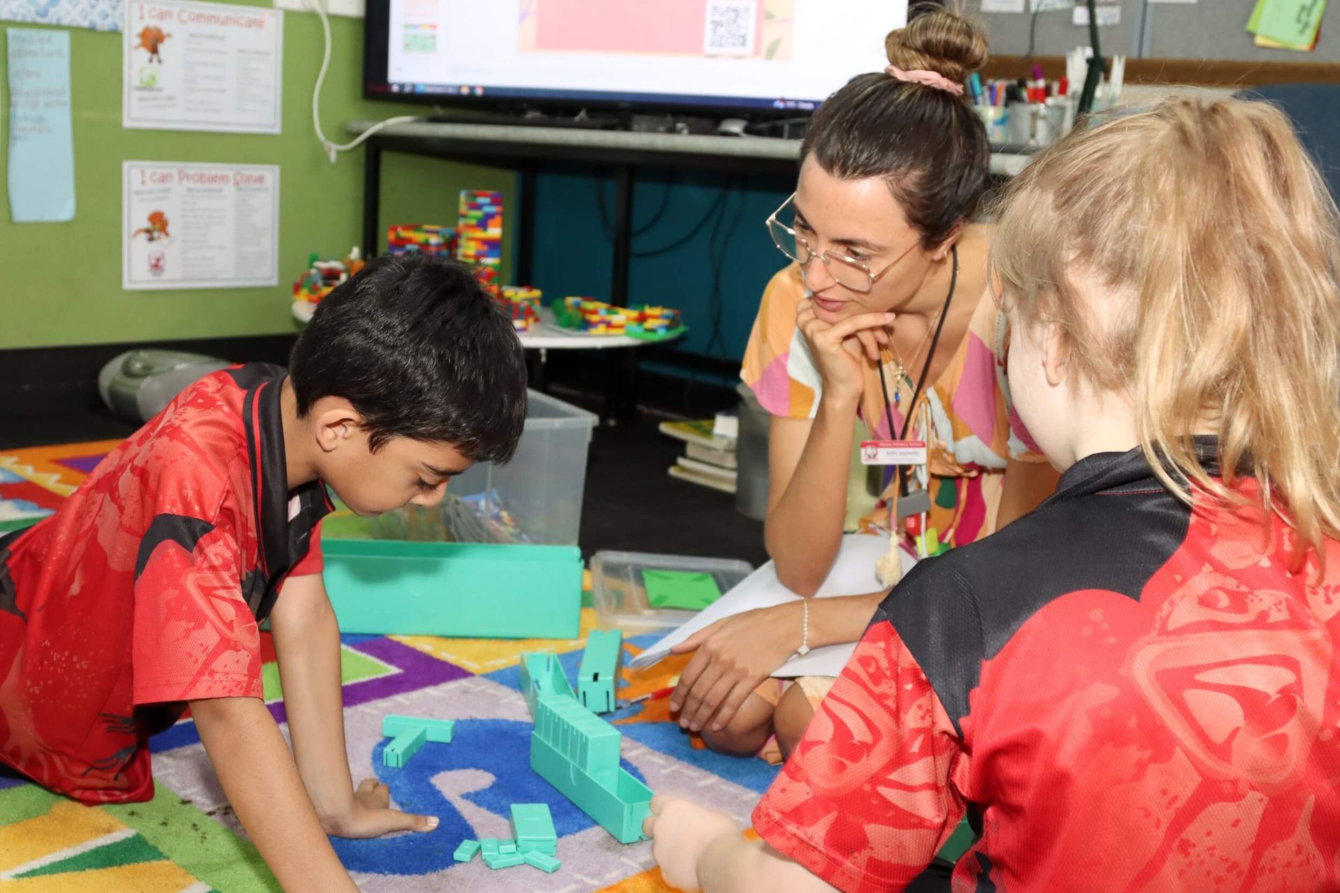 Teacher assisting two students with a building activity, using colored blocks, in a classroom setting.