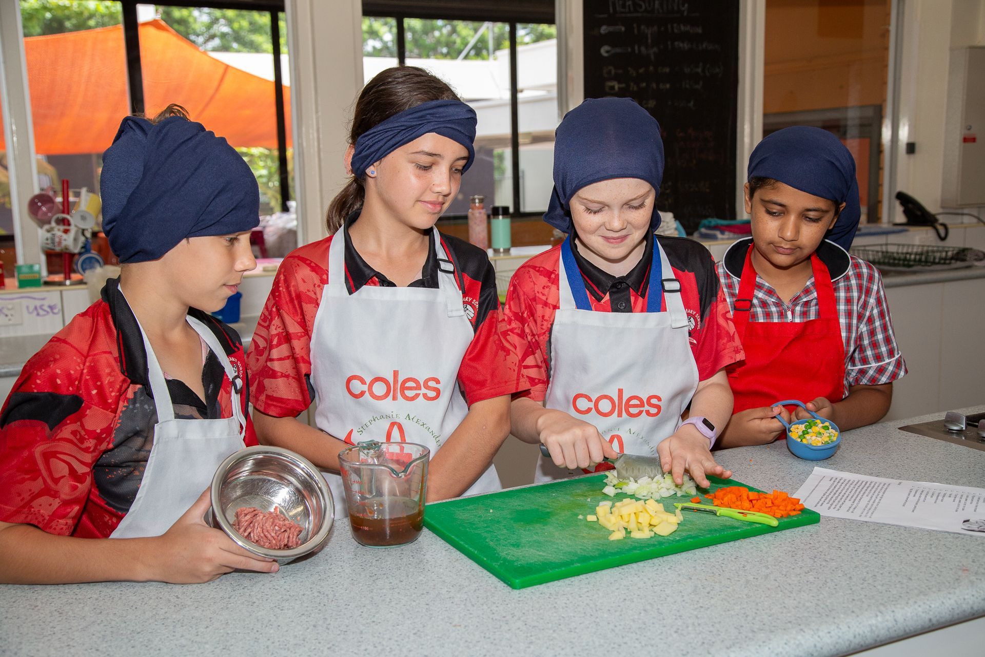 Four children in aprons and head coverings, chopping vegetables in a kitchen.