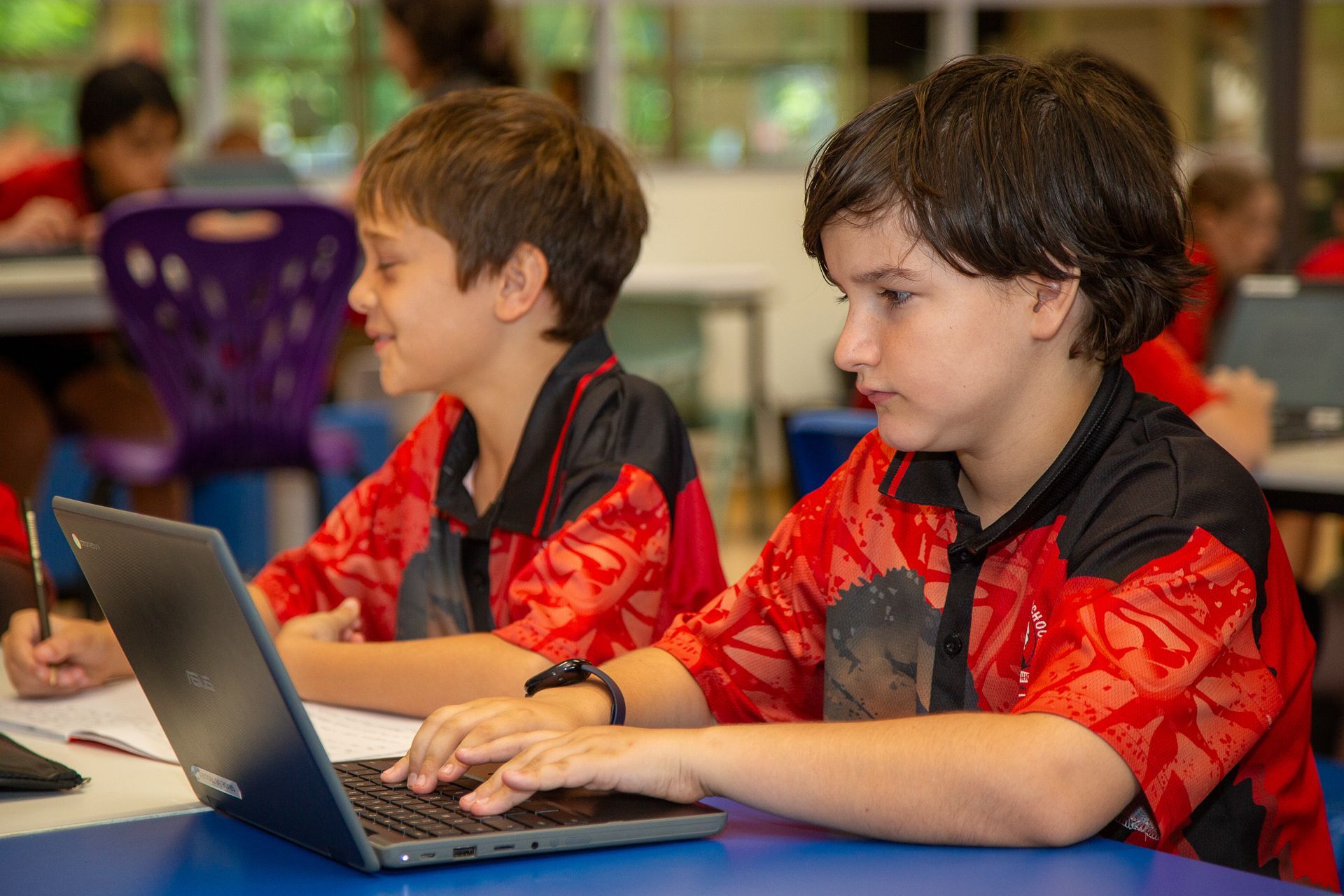 Two students in red shirts work on a laptop in a classroom.