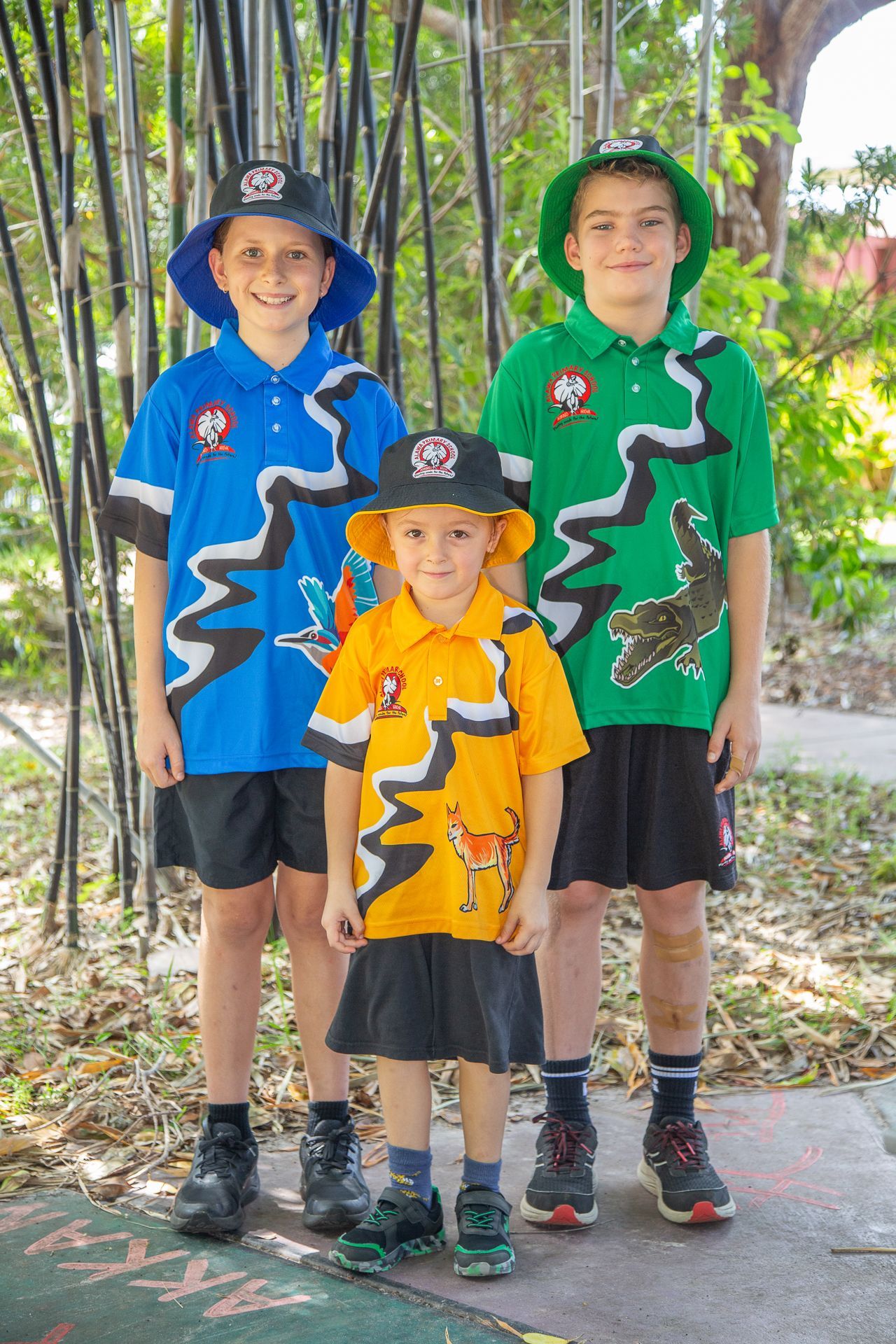 Three children in school uniforms stand outdoors; blue, green, and orange shirts with matching hats.