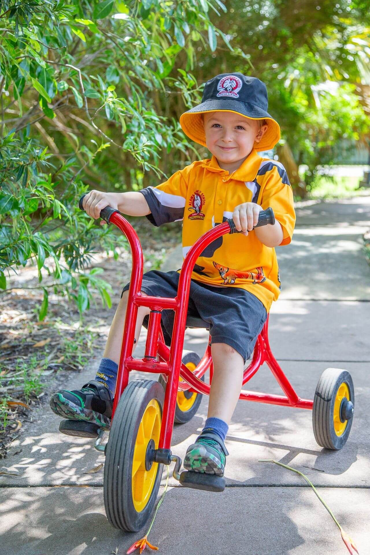 Boy smiling, riding a red tricycle outdoors, wearing a yellow shirt and a sun hat.