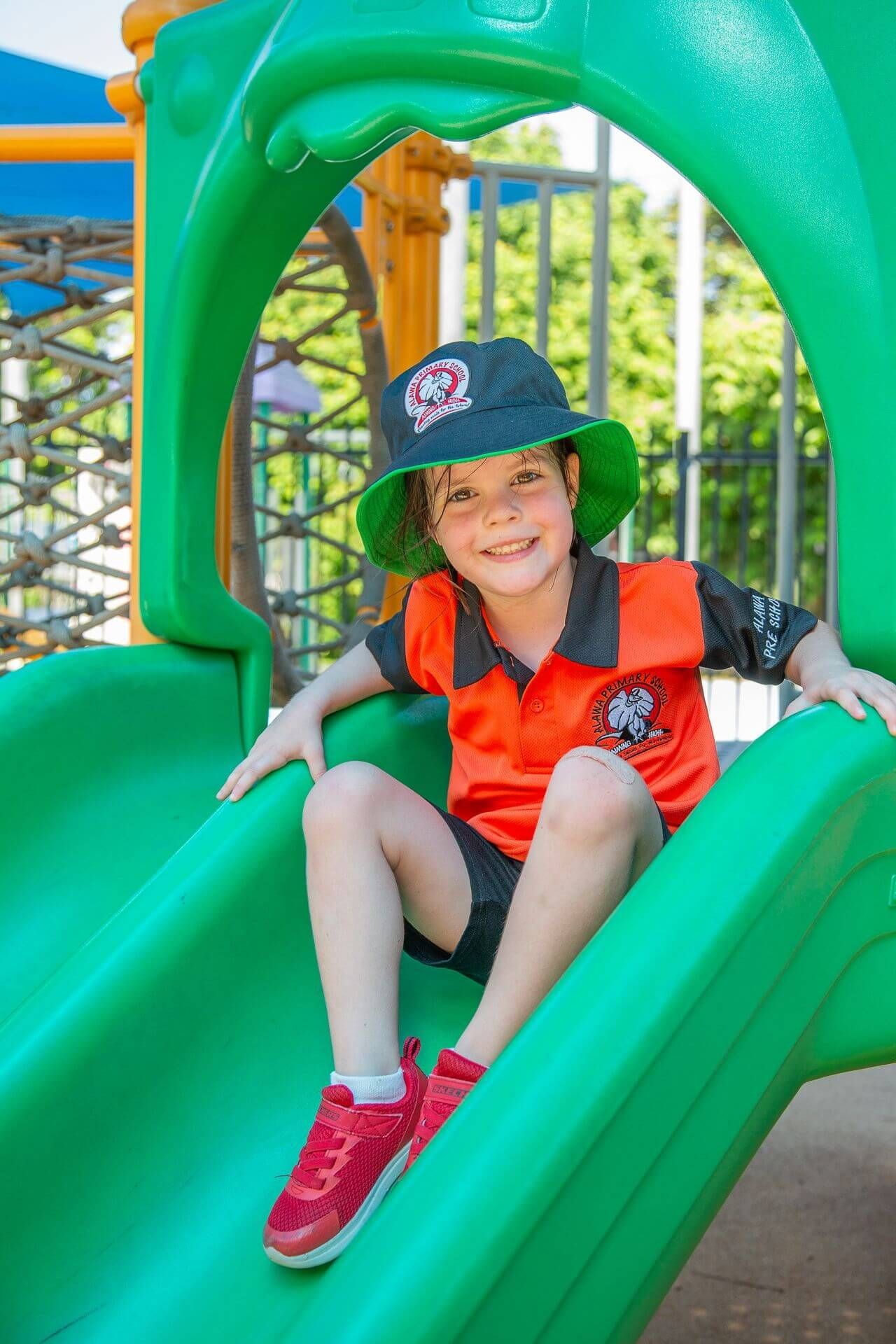 Child wearing school uniform smiles on a green slide at a playground.