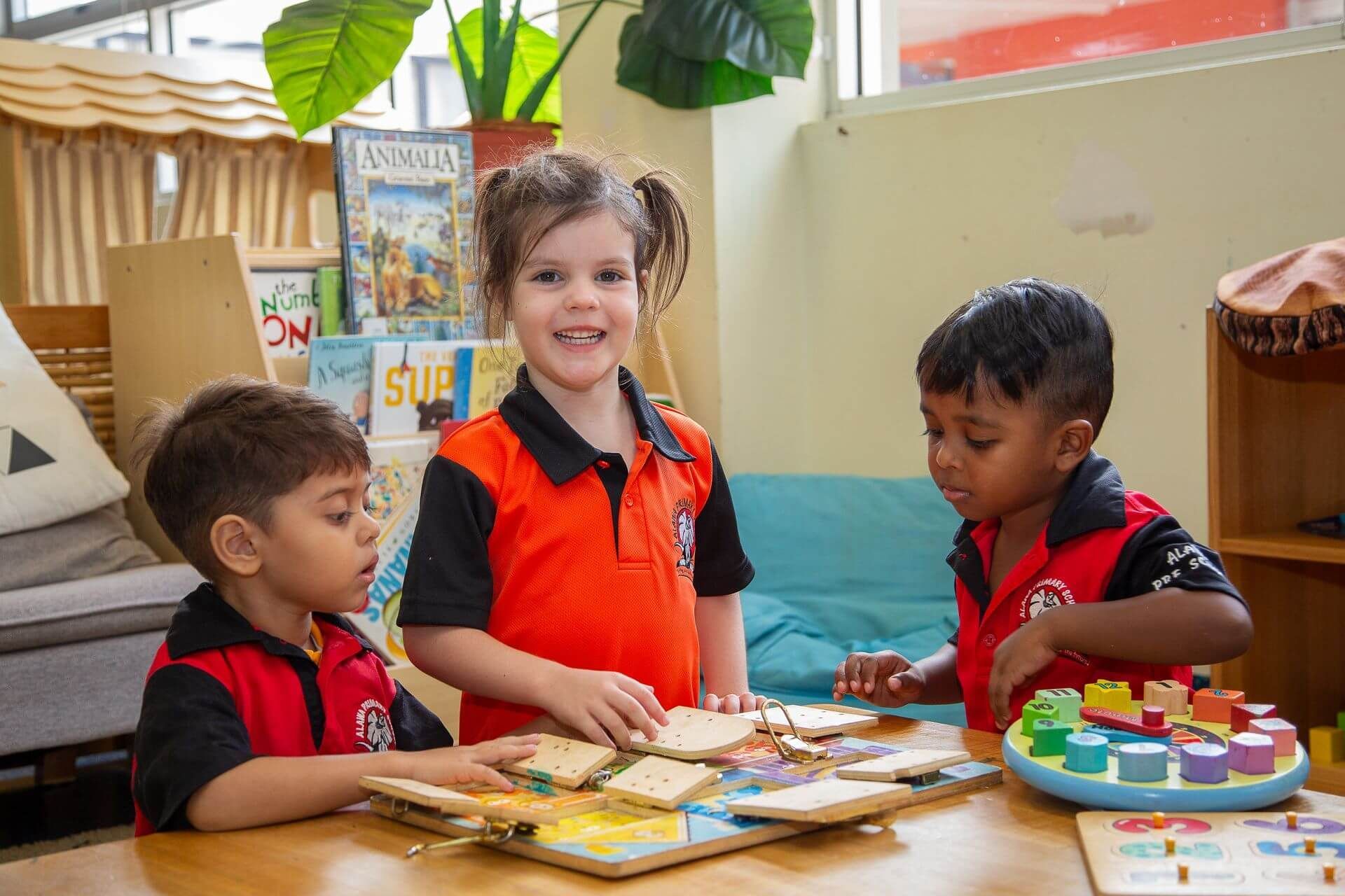 Three children in orange shirts playing with puzzles at a table indoors.