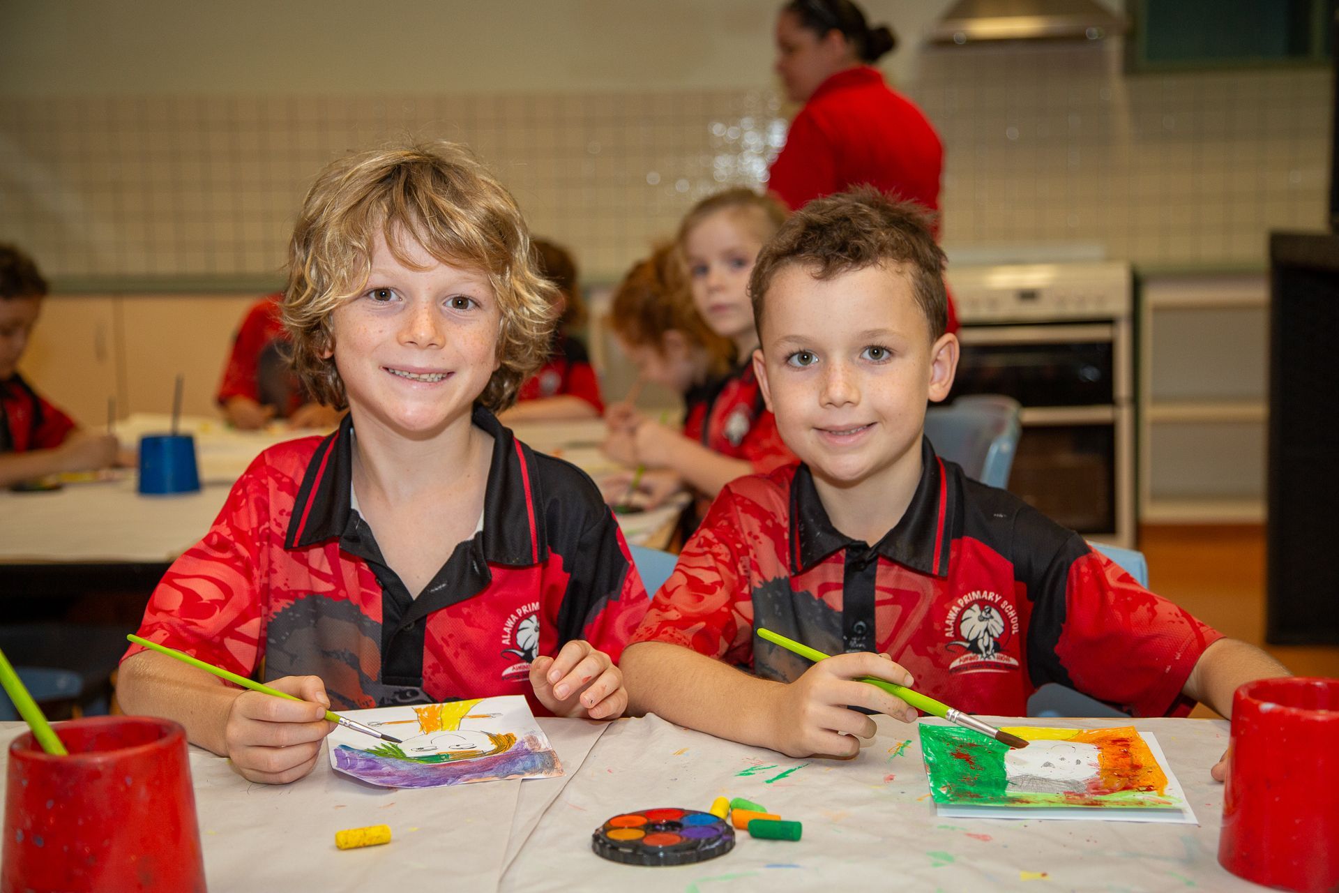 Two boys in red shirts painting at a table in a classroom.