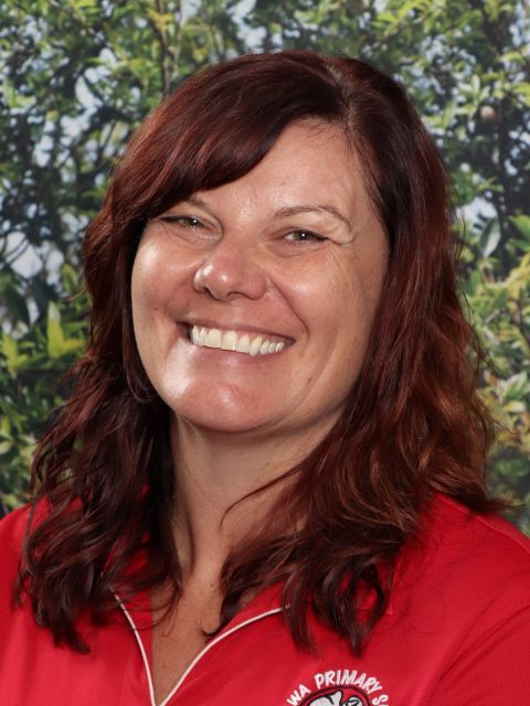 Woman with auburn hair smiles, wearing a red top, in front of greenery; likely a school setting.