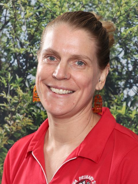 Woman with blonde hair in a red polo shirt smiles; background is green foliage.