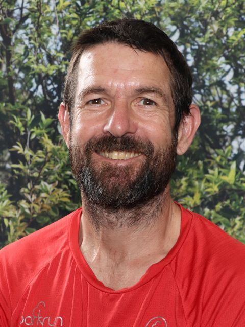 Man with a beard, smiling, wearing a red shirt, in front of a green leafy background.
