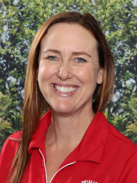 Woman smiling, wearing a red shirt, in front of a green leafy background.