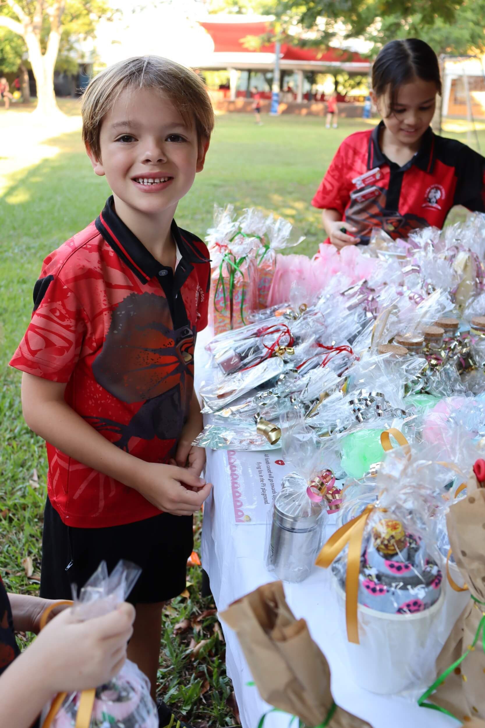 Boy smiling at a table of wrapped gifts, another person in the background. Red shirts, outdoor setting.