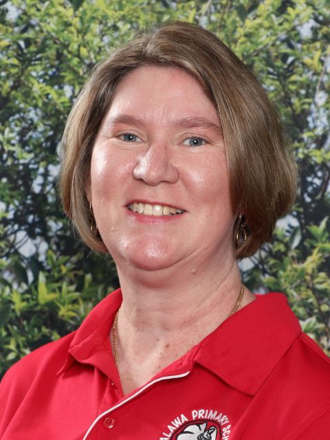 Woman with short brown hair in a red shirt, smiling, set against green foliage.