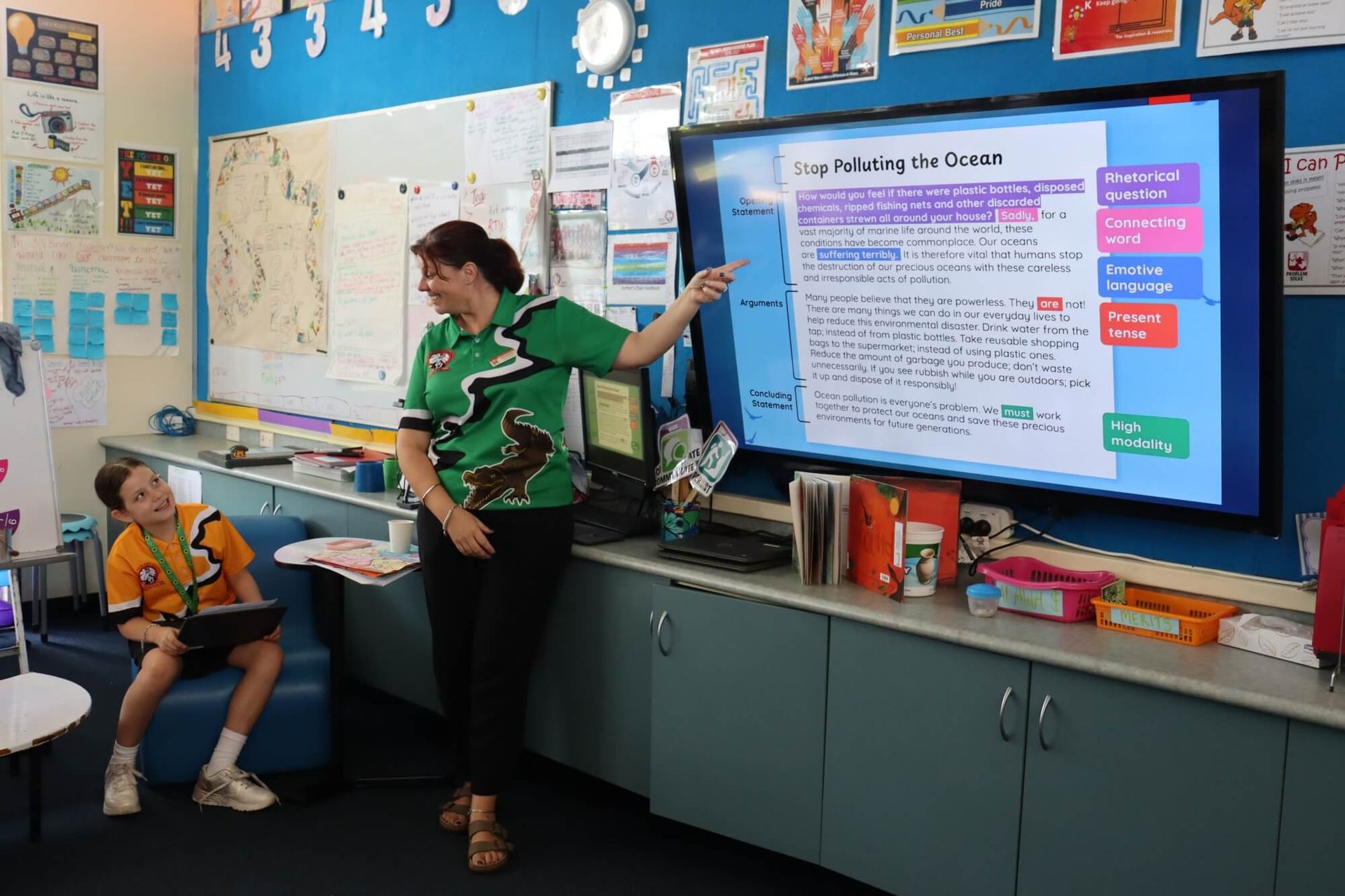 A teacher points at a screen in a classroom, with a student seated nearby.