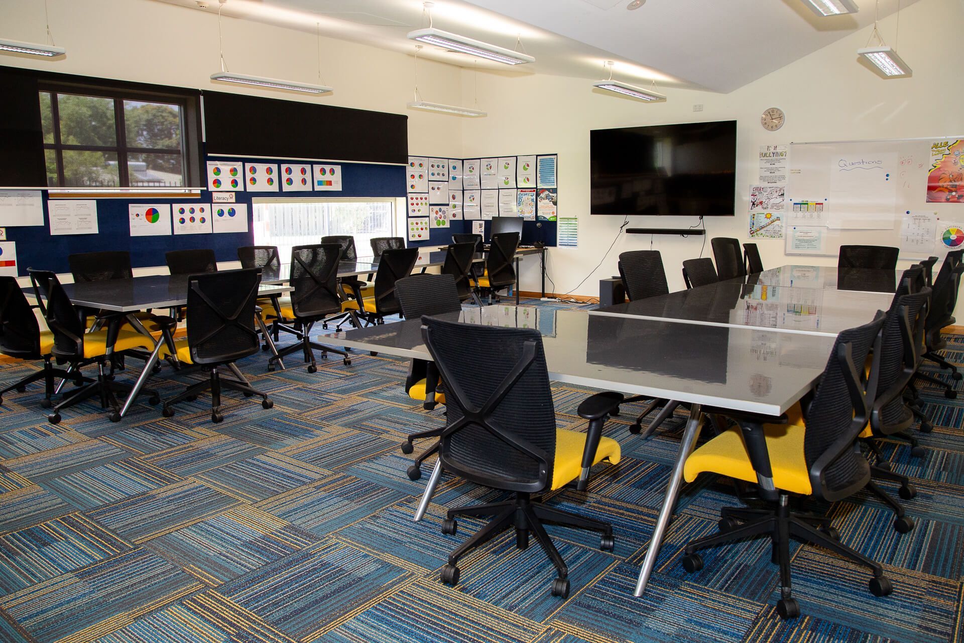 Classroom with long tables, black and yellow chairs, a large screen, and work displayed on the walls.