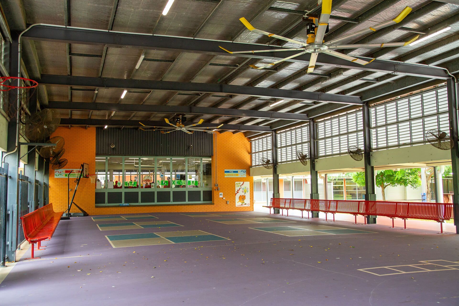 Indoor gymnasium with purple floor, orange walls, red benches, and ceiling fans.