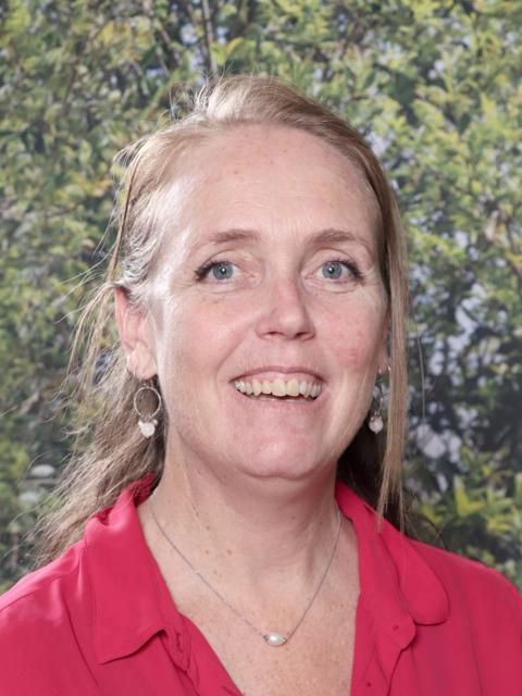 Woman with auburn hair smiles, wearing a red top, in front of greenery; likely a school setting.