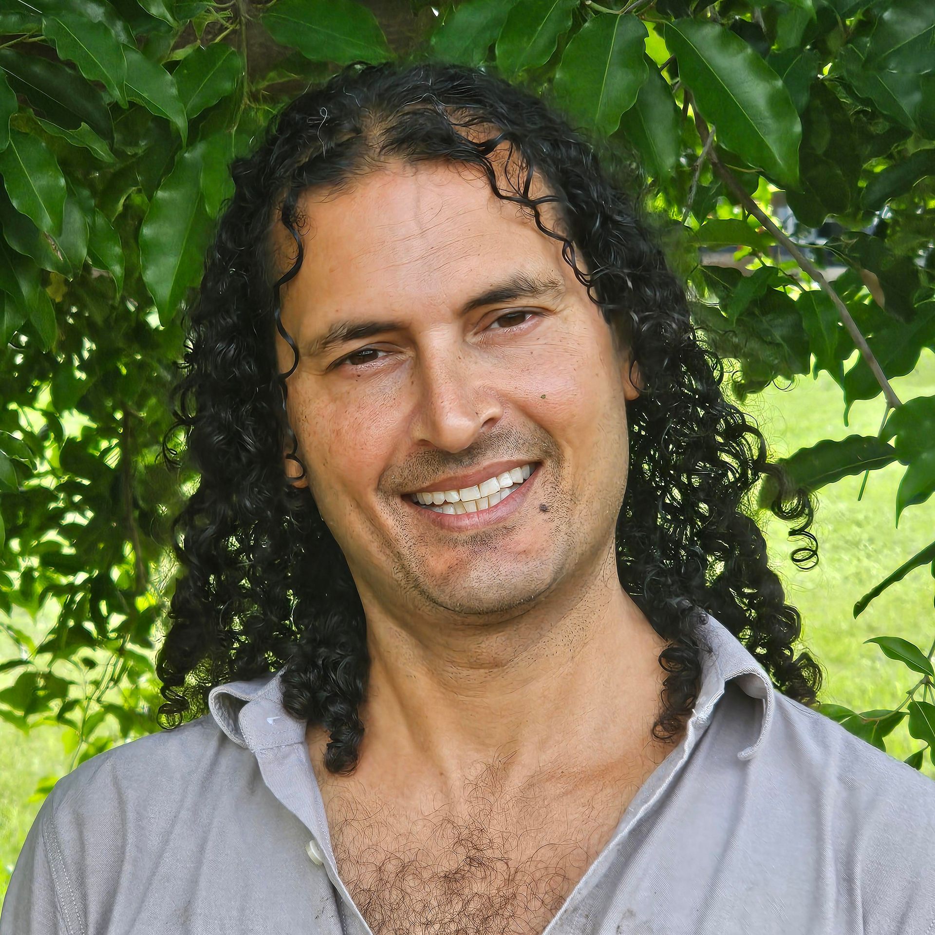 Man with a beard, smiling, wearing a red shirt, in front of a green leafy background.