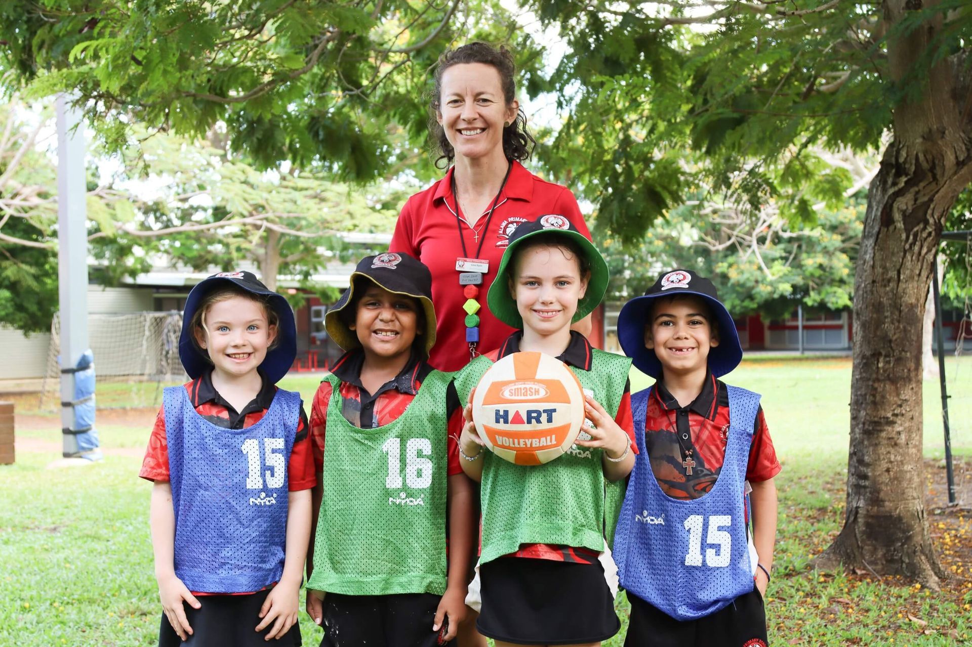 Woman and four children in netball uniforms holding a ball. Outdoors, trees in background.