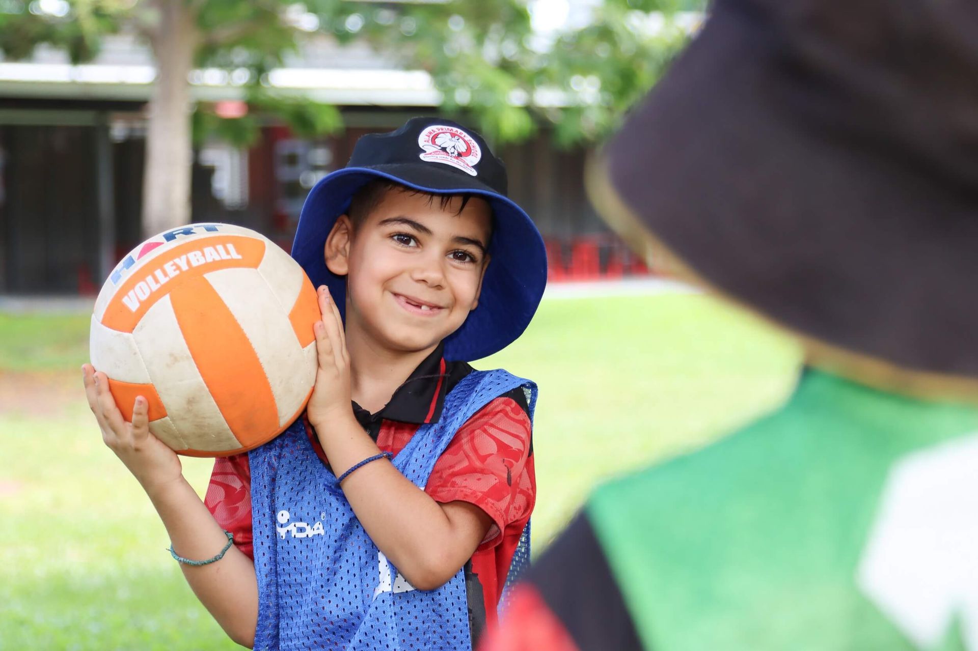 Boy holding volleyball, smiling, wearing hat and vest on a green field.