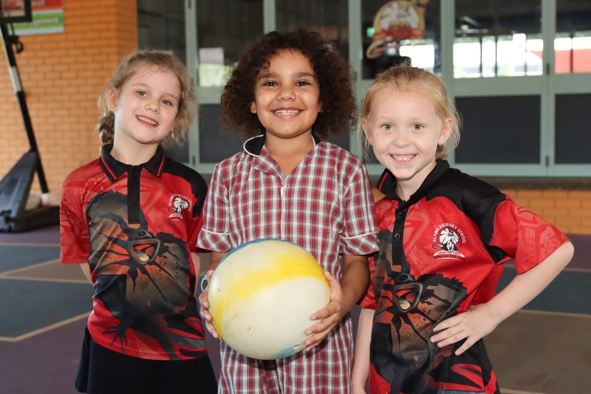 Three children smiling, one holding a ball. Two wearing red jerseys, all stand near a building.