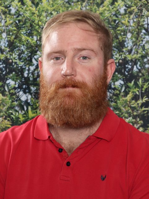 Man with red beard and shirt, neutral expression, in front of green foliage.