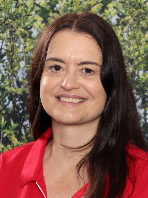 Woman with long dark hair smiling, wearing a red shirt against a green leafy background.