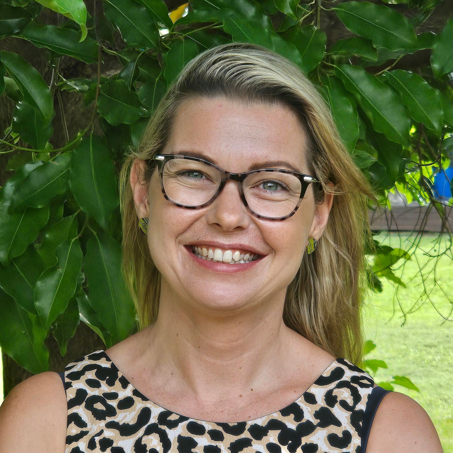 Woman with brown hair, smiling in a red shirt, against a green leafy background.