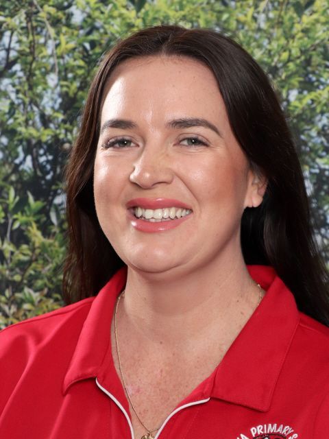 Woman with dark hair and red shirt smiling, in front of green foliage.