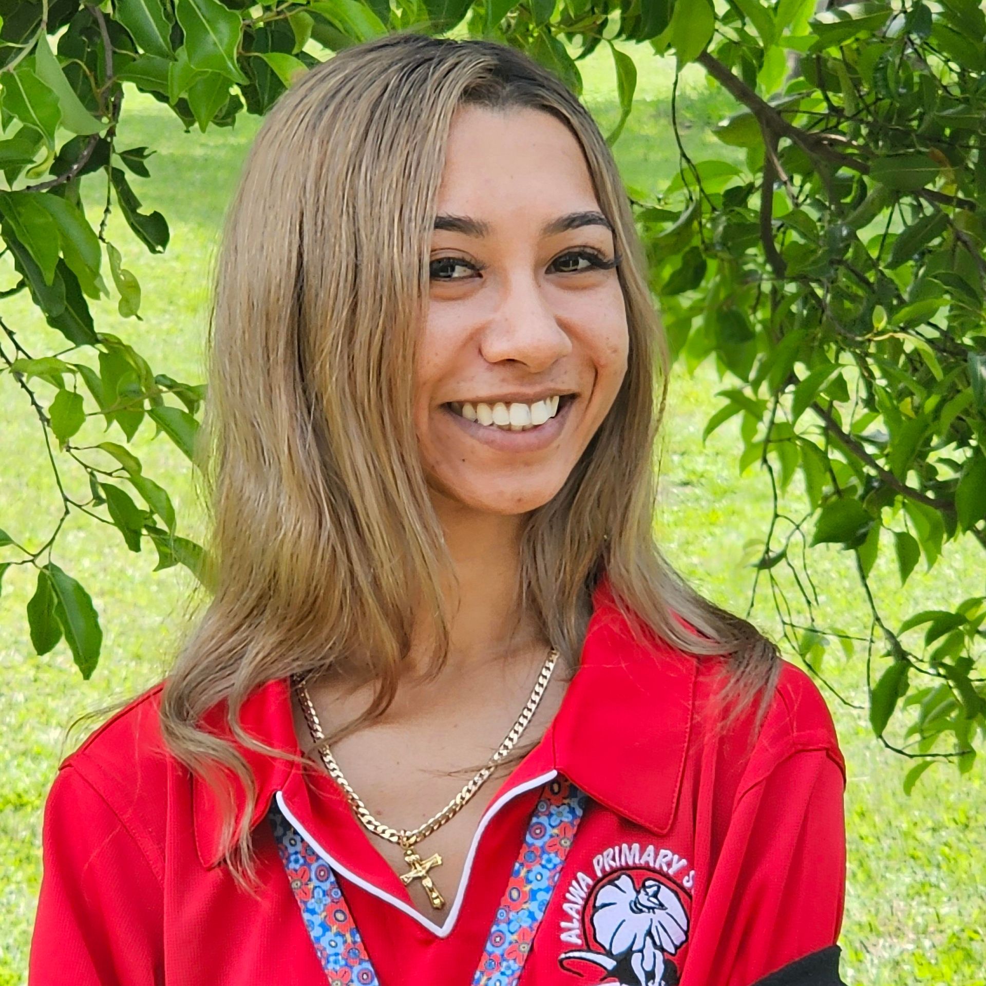 Woman in a red polo shirt with a logo smiles in front of a green foliage background.