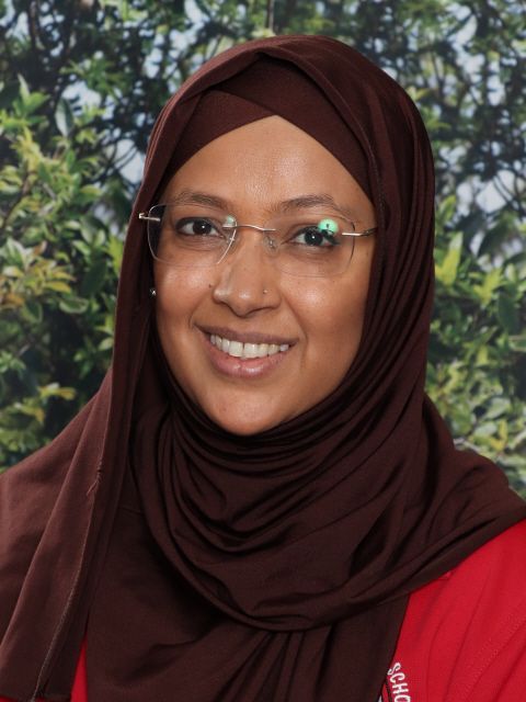 Woman wearing glasses and a brown hijab, smiling in front of greenery.